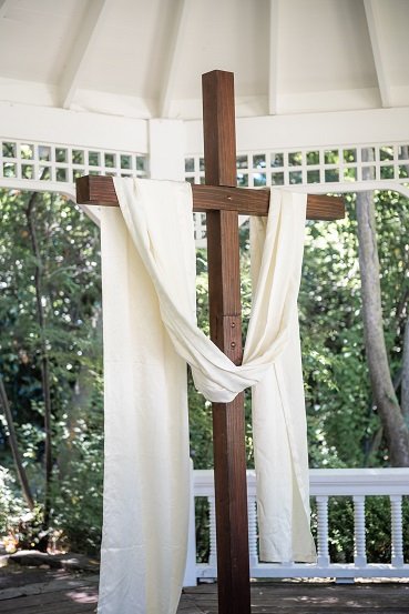 A wooden cross draped with white cloth hangs against a neutral background.