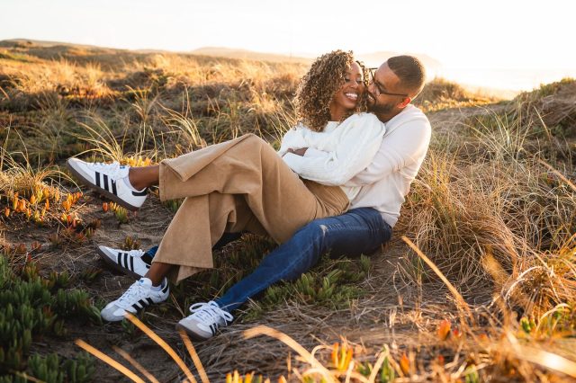 A man and woman are sitting together in a grassy field near a beach.